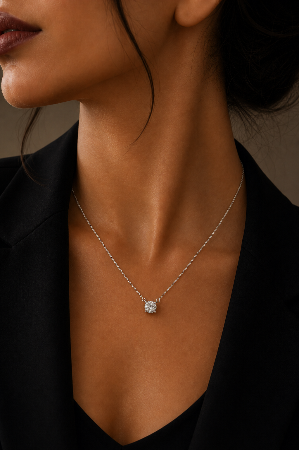 A close-up of a model wearing a delicate silver chain necklace with a single round clear gemstone pendant, styled with a black blazer.