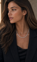 A close-up of a woman with long brown hair wearing a delicate silver chain necklace, styled with a black blazer and black top.