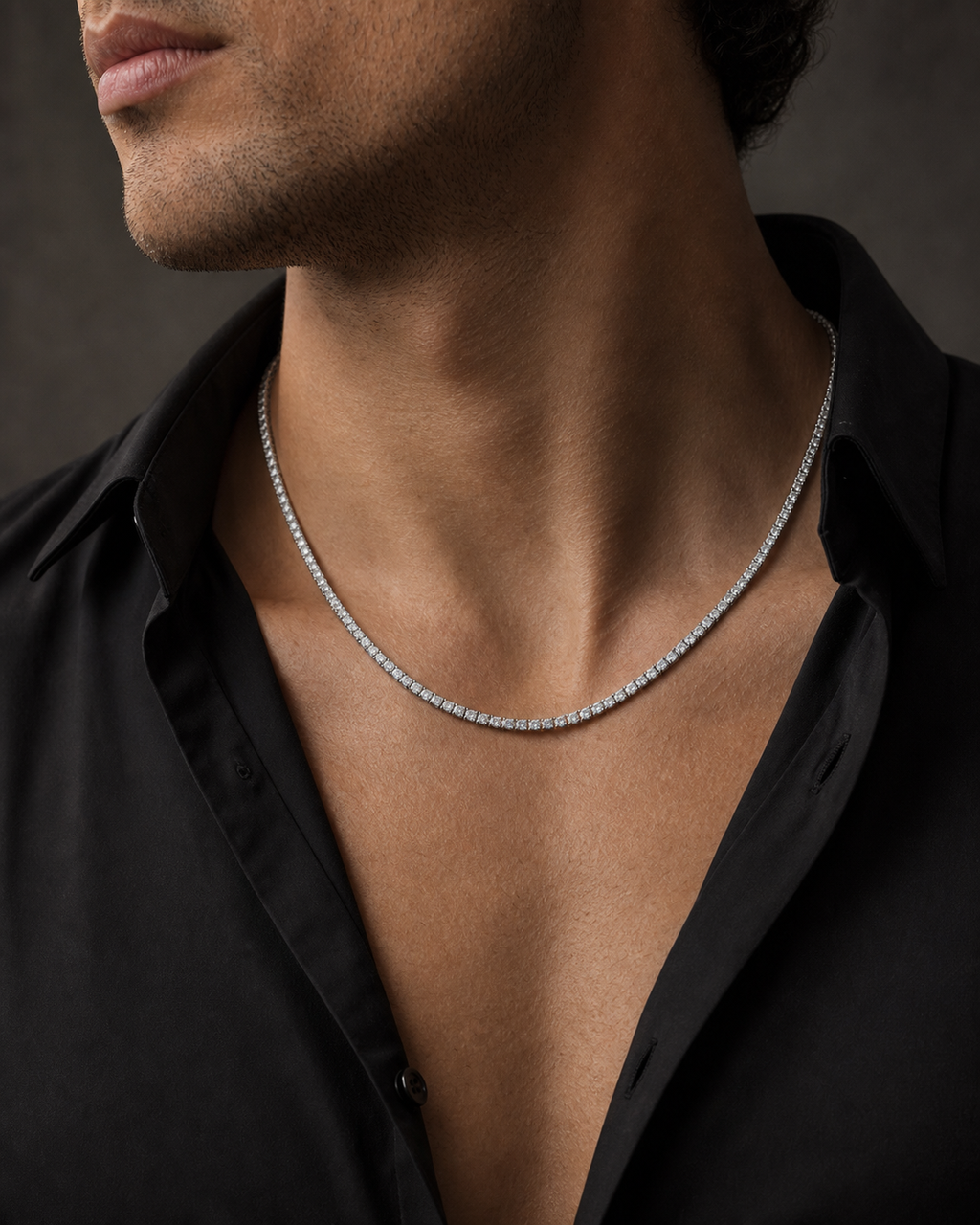 A close-up of a man's neck and chest wearing a delicate silver tennis chain necklace over a black button-down shirt.