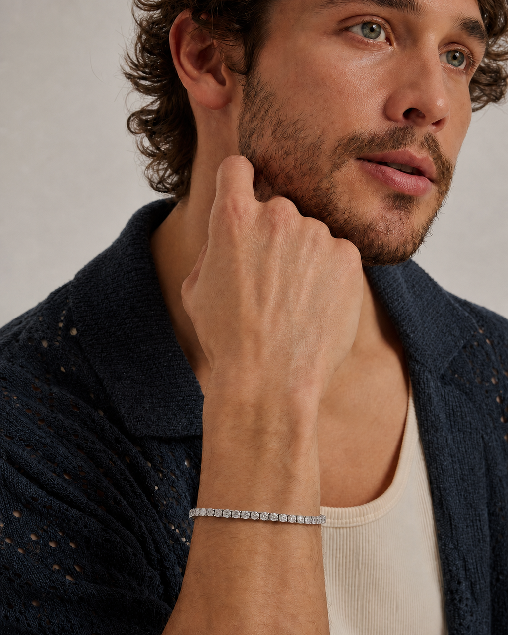 A close-up of a man wearing a delicate silver tennis bracelet, styled with a dark textured knit cardigan and a cream ribbed tank top against a neutral light gray background.