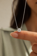 A close-up of a delicate silver chain necklace with a single round-cut clear gemstone pendant, held between a person's fingers against a softly blurred background.