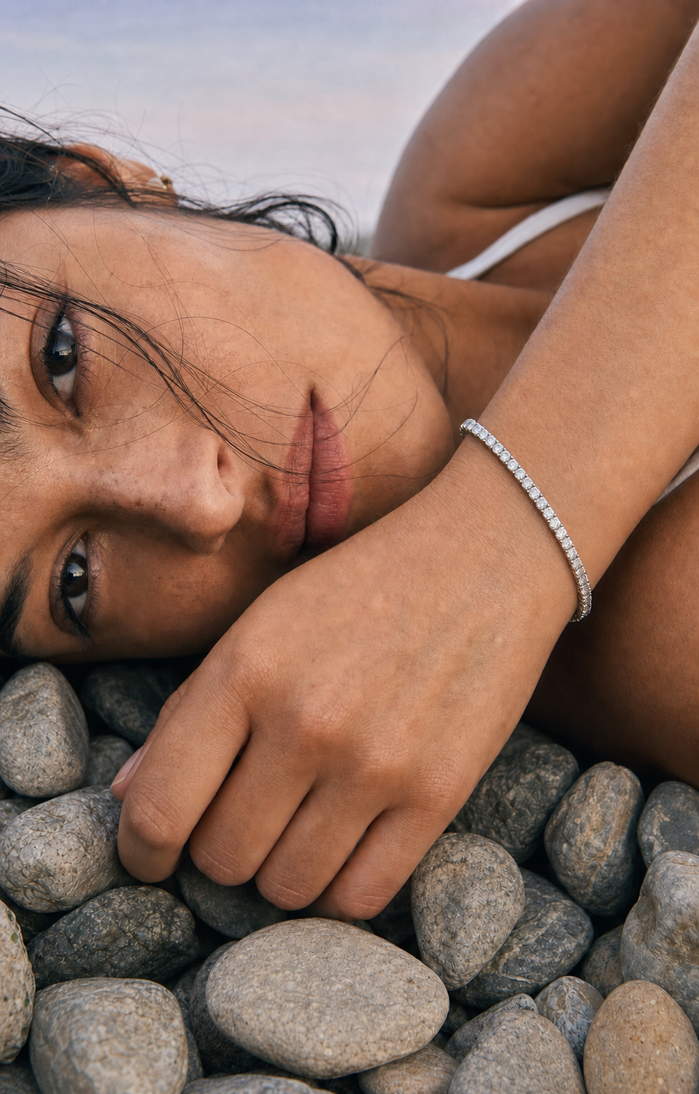 A close-up lifestyle shot of a woman lying on smooth gray pebbles at the beach, wearing a delicate silver chain bracelet on her wrist.